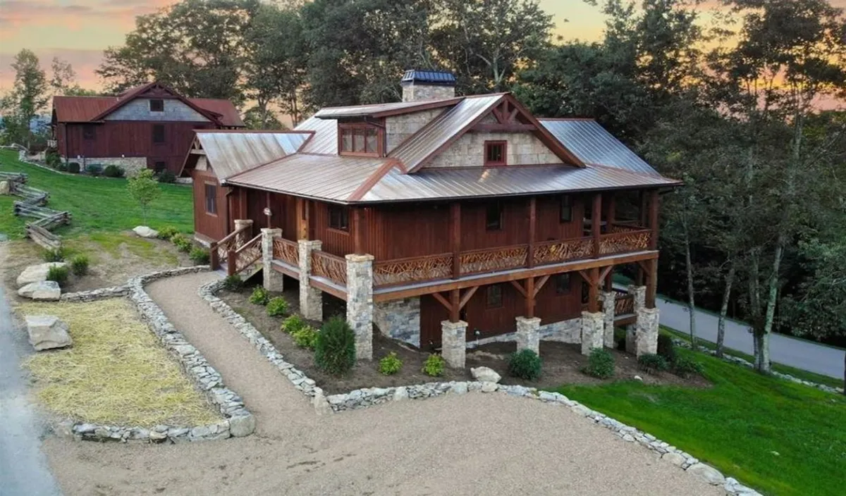 The image showcases the exterior of Timberwolf Lodge, a striking wooden cabin with stone accents, surrounded by lush landscaping. The scene invites guests to enjoy the expansive balcony with sunset mountain views, a welcoming fire pit area, and an atmosphere perfect for family gatherings or relaxing outdoor evenings.