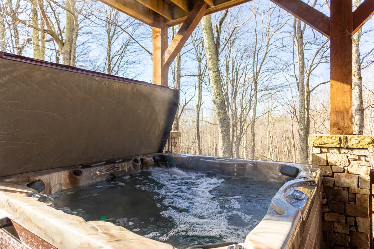 A tranquil view of an outdoor hot tub nestled beside a wooden structure, surrounded by tall trees. The setting feels private and immersed in nature, perfect for soaking in the fresh mountain air while enjoying the forest views at sunrise or under the stars.