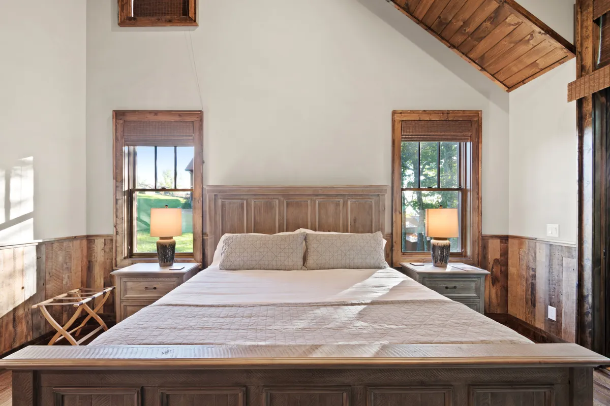 A tranquil bedroom featuring a wooden bed frame with decorative pillows, table lamps on each side, and windows that let in soft natural light. The space is styled for comfort and relaxation, suggesting a peaceful retreat.