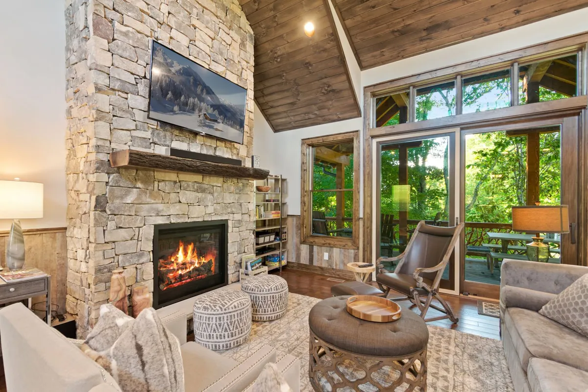 A cozy living room featuring a stone fireplace, plush seating, decorative poufs, and large windows that offer sunset views. The wooden ceiling contributes to the mountain lodge feel, while the arrangement encourages relaxation and intimate conversation.
