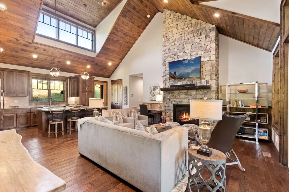 A spacious open-plan area featuring a stone fireplace flanked by comfortable seating and large windows that let in ample natural light. The kitchen island is visible in the background, seamlessly connecting the living and kitchen spaces. The overall atmosphere is inviting, with warm lighting and thoughtful decor.