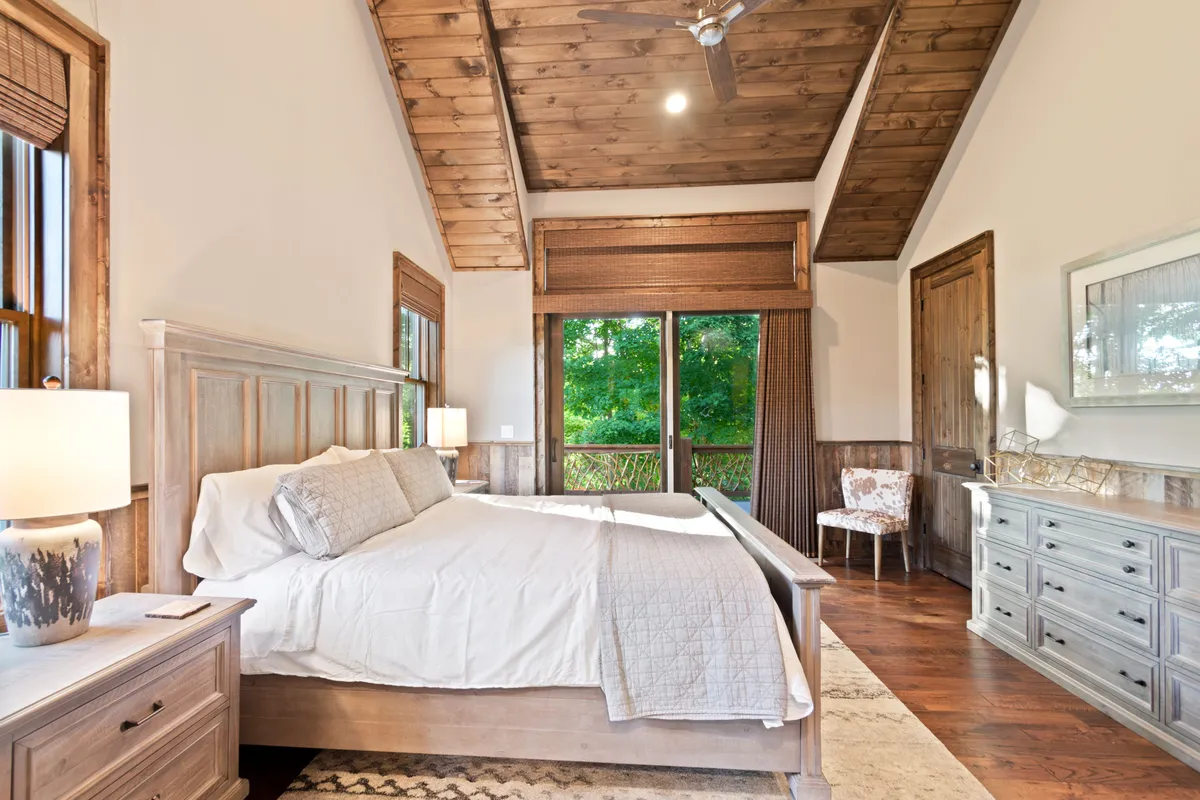 A calm, cozy bedroom with a wooden bed frame, large windows offering views of nature, a decorative chair, and a dresser. The room feels inviting, serene, and designed for relaxation.