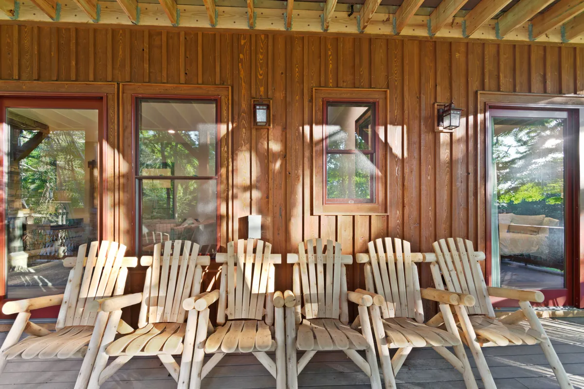 A cozy exterior scene featuring wooden chairs arranged along a wooden wall under outdoor light fixtures, windows framing the view, and gentle sunlight illuminating the space. The setup invites moments of relaxation and socializing while enjoying the peacefulness of the surrounding environment.