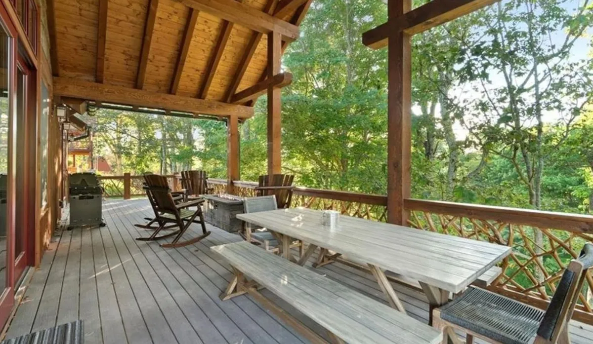 An exterior view focused on a spacious wooden deck with comfortable outdoor seating and a dining table, surrounded by lush greenery. The setting is inviting, ideal for barbecuing, family gatherings, or simply sitting and enjoying the peaceful natural surroundings.