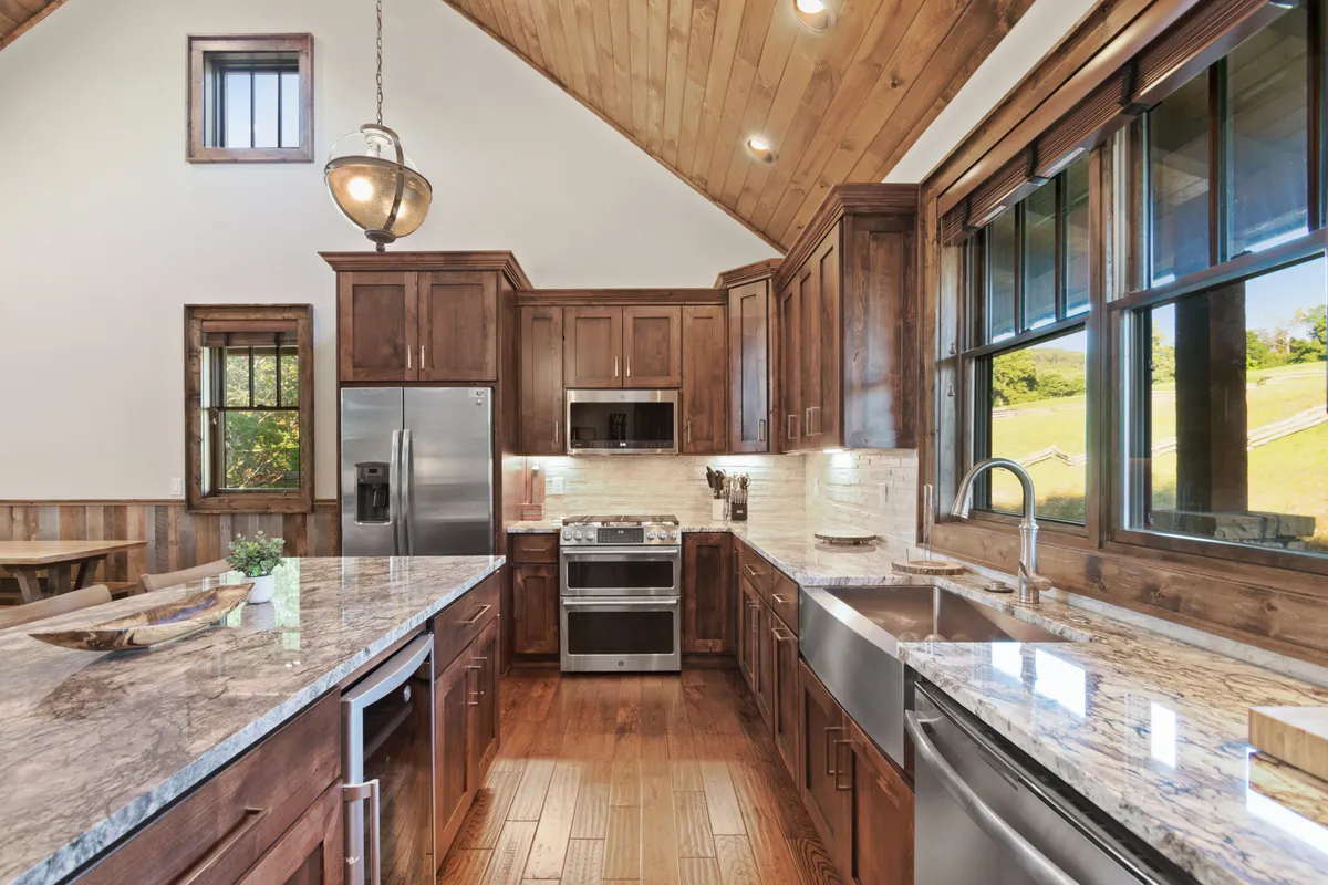 A close-up of a well-equipped kitchen with granite countertops, wooden cabinetry, stainless steel appliances, and large windows that provide natural light and glimpses of the outdoors. There is ample space for cooking, entertaining, and gathering as a group.